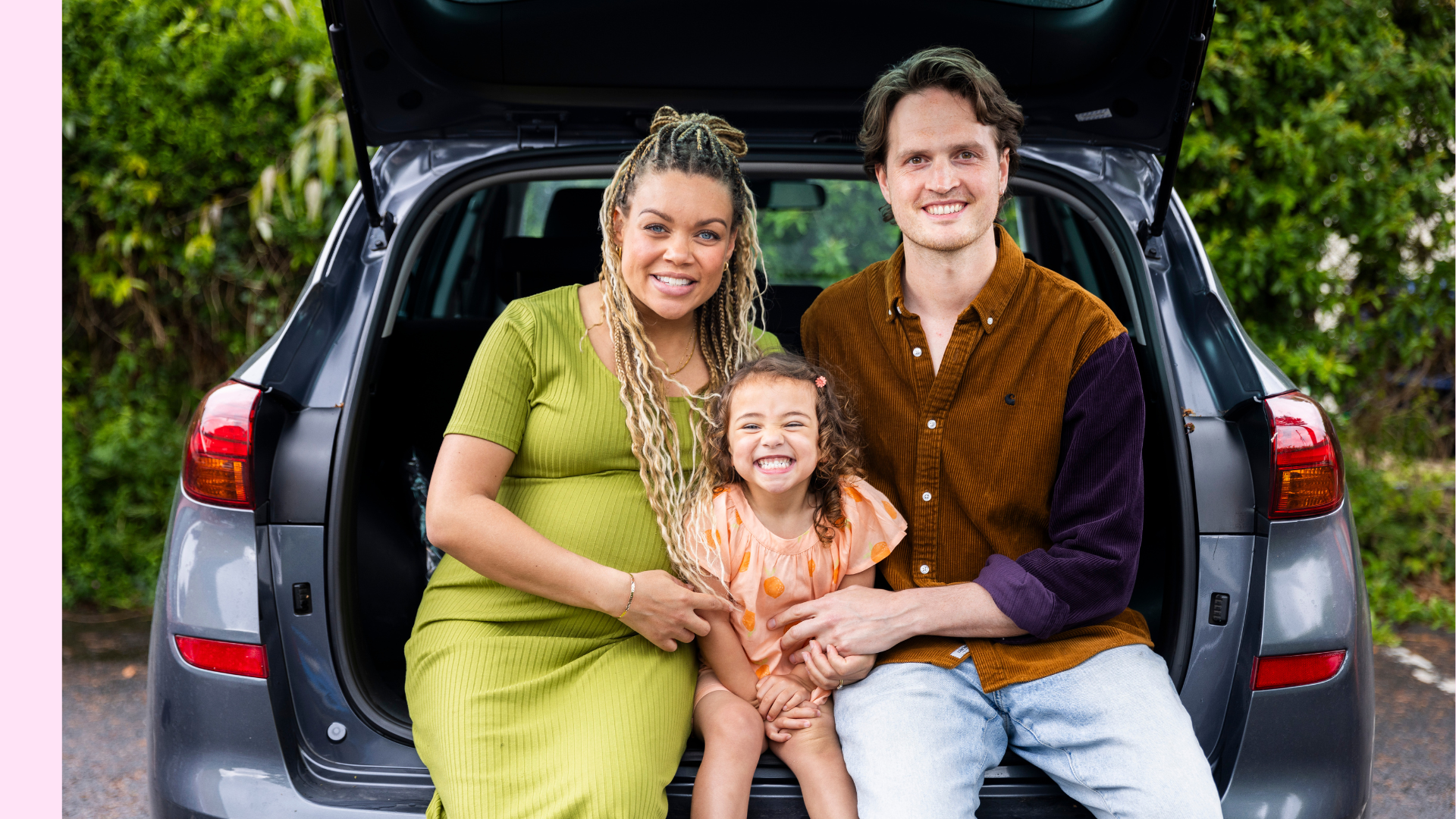 Couple sat in the boot of a large car with daughter in the middle of them, all three are smiling and looking at the camera